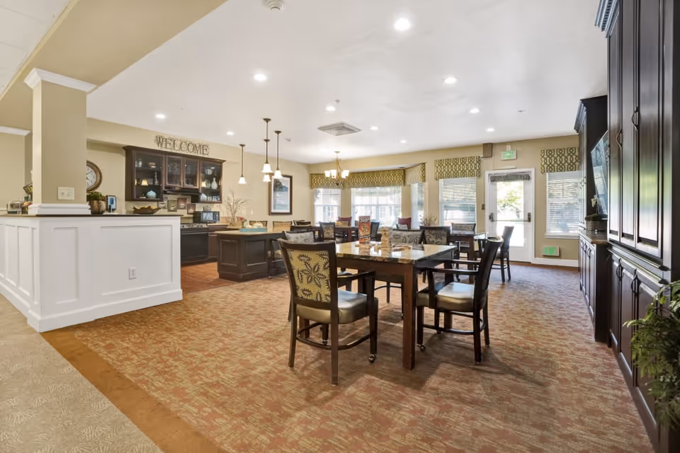 A spacious and well-lit dining area in a senior living facility named Dale Commons, featuring multiple wooden tables and chairs with patterned upholstery. The room has large windows with green patterned valances allowing natural light to fill the space. There is a kitchen area with dark cabinetry, a countertop island, and pendant lighting. The walls are painted in neutral tones, and a 'WELCOME' sign is displayed above the kitchen cabinets.