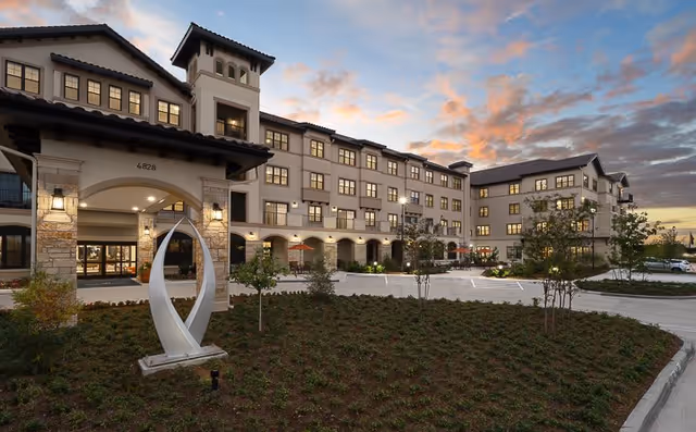 Exterior view of a large, multi-story senior living facility building at sunset with well-maintained landscaping, a modern sculpture near the entrance, and a parking area in front.