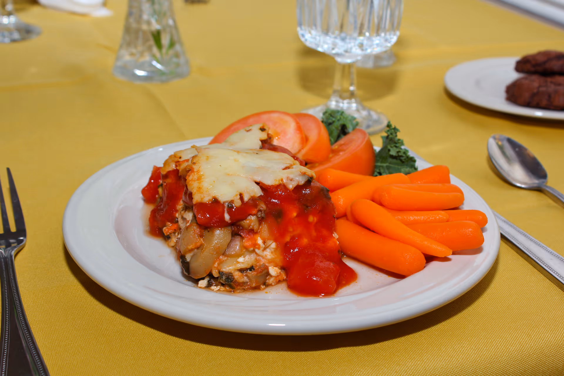 A plated meal of cheesy casserole with tomato sauce, baby carrots and sliced tomatoes on a yellow tablecloth with a water goblet and utensils.
