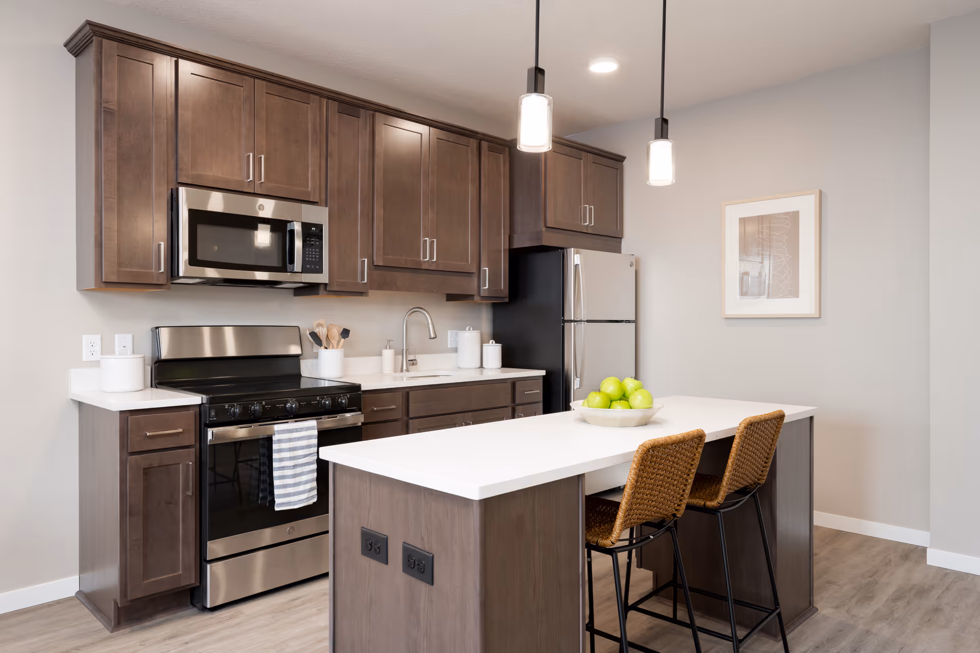 Modern kitchen with dark wood cabinets, stainless steel appliances including a microwave, stove, and refrigerator. A white countertop island with two wicker bar stools and a bowl of green apples sits in the center. Two pendant lights hang above the island, and a framed artwork is on the wall.