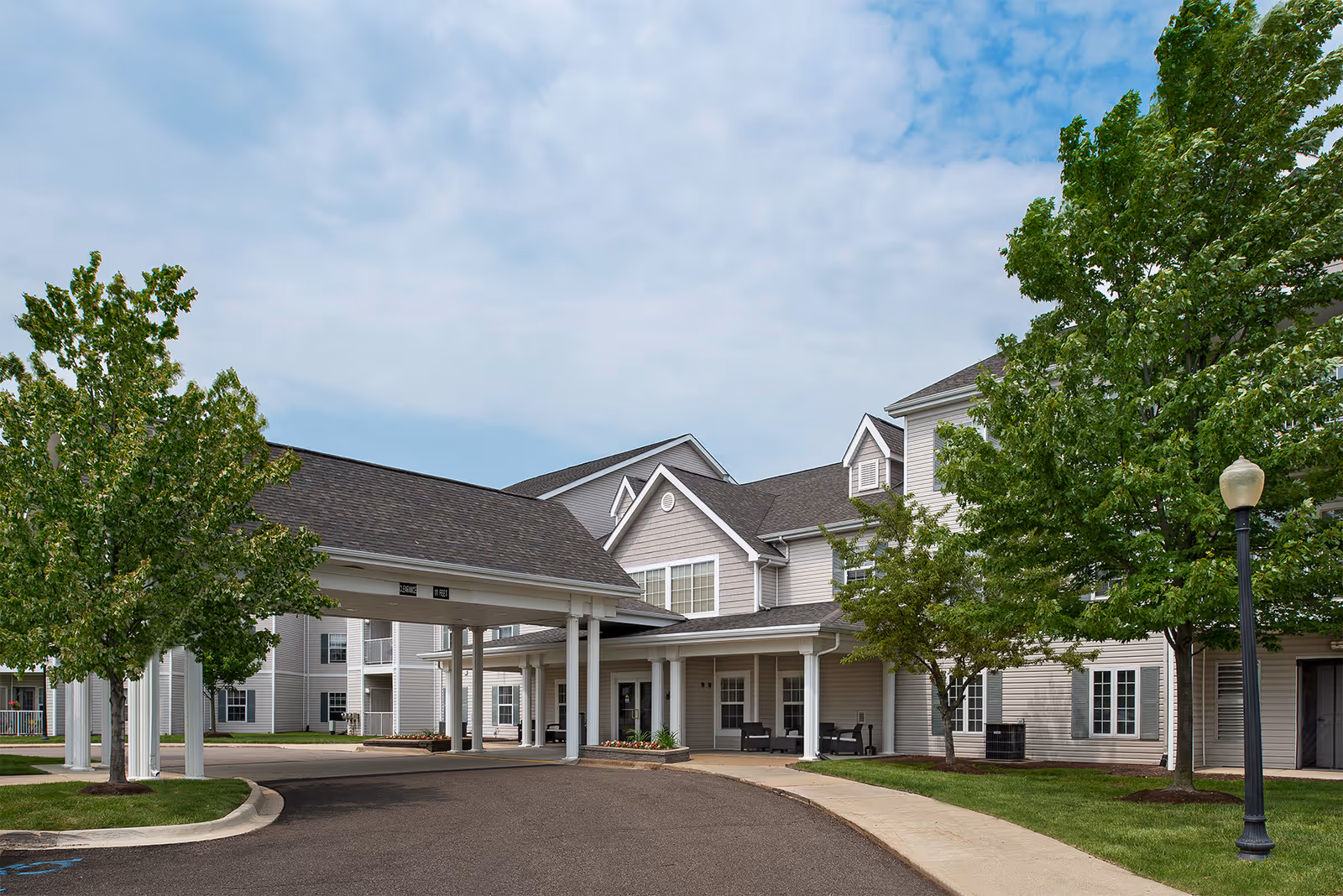 Exterior view of American House Troy senior living facility showing a covered entrance with white pillars, a paved driveway, green trees, and a multi-story building with gray siding and white trim under a partly cloudy sky.