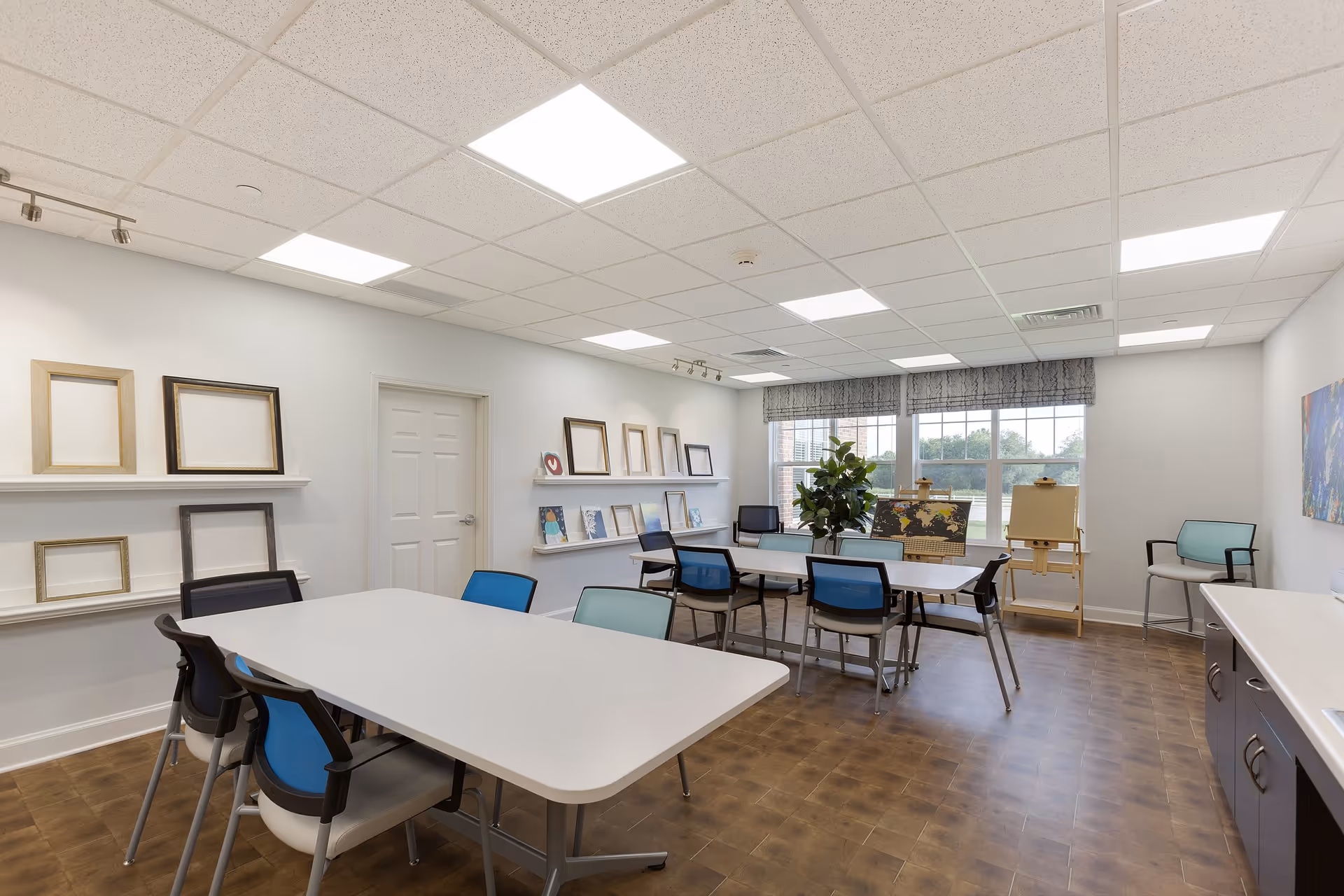 A bright, spacious activity room with two white tables surrounded by chairs with blue and light blue backs. The room has wooden flooring, white walls, and a drop ceiling with recessed lighting. On the left wall, there are shelves displaying empty picture frames and some small artworks. At the far end, large windows with patterned valances let in natural light. There is a plant and two easels near the windows, one holding a world map. A countertop with cabinets is visible on the right side of the image.
