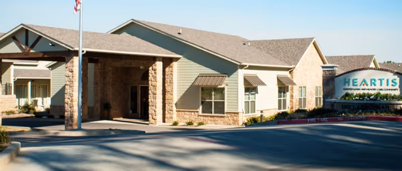 Exterior view of Heartis Longview senior living facility showing the main entrance with a covered drop-off area, stone and siding facade, windows with awnings, and a sign with the facility name.