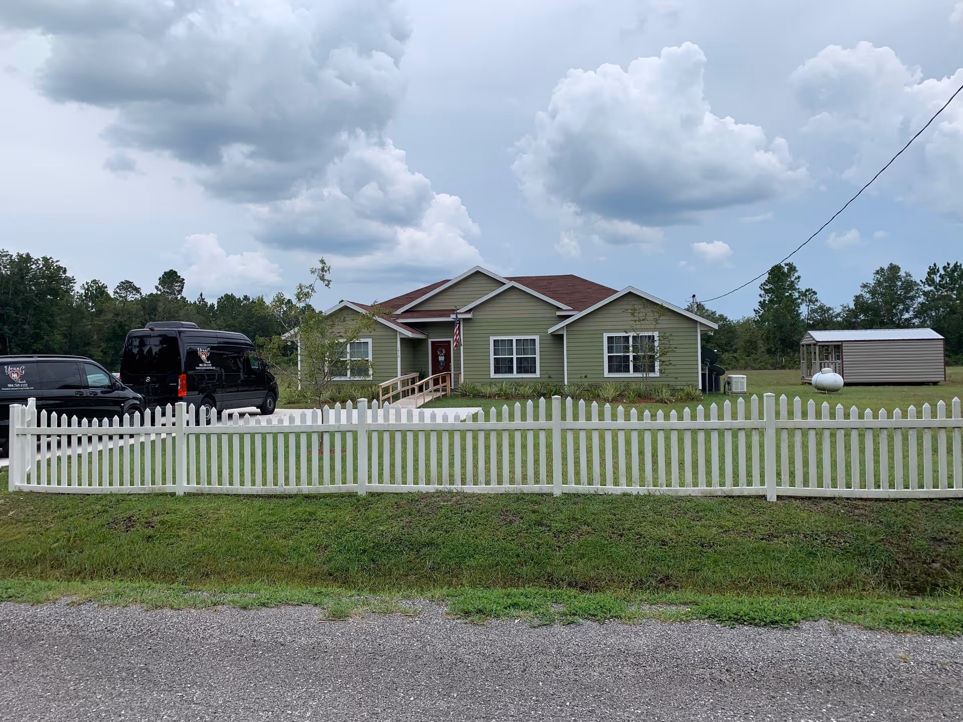 Single-story green house with a white picket fence, driveway with black vans, a small shed, and a cloudy sky.