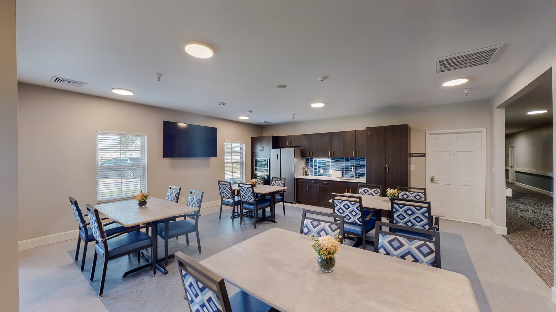 A bright and modern communal dining area with several tables and chairs featuring blue and white patterned upholstery. The room includes a kitchen area with dark wood cabinets, a refrigerator, an oven, and a blue tiled backsplash. There is a large flat-screen TV mounted on the wall between two windows with blinds. The floor is a combination of light and darker tones, and the ceiling has multiple round recessed lights.