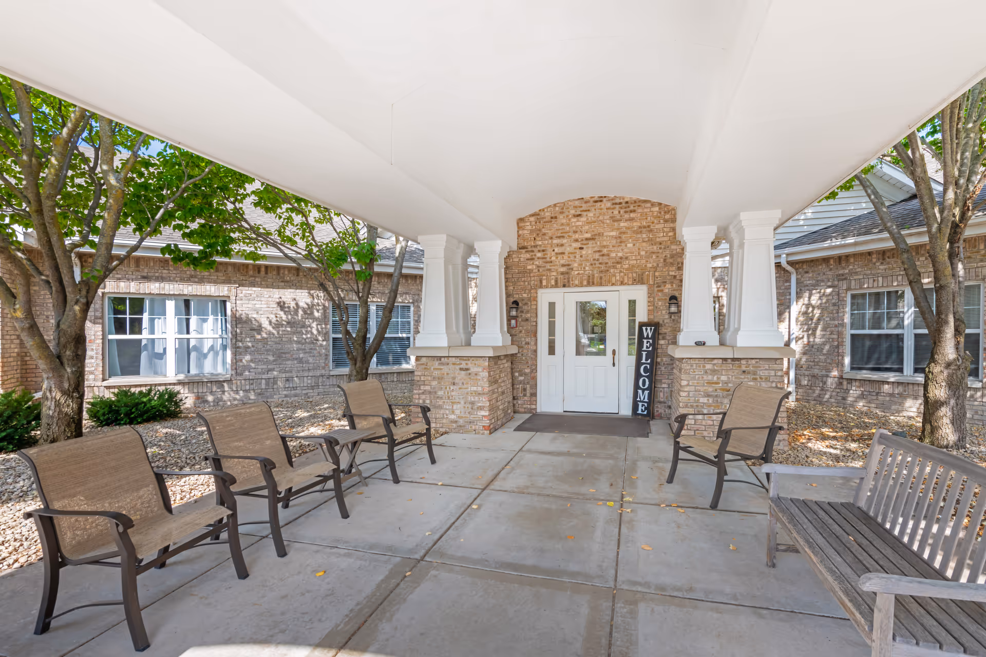 Covered brick entrance with chairs and a 'Welcome' sign at the front door of the senior living facility.