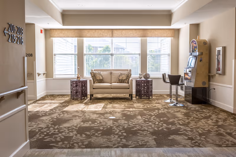 A bright sitting area in a senior living facility with a beige loveseat flanked by two decorative side tables, large windows with blinds letting in natural light, a slot machine with two bar stools on the right, and directional signs on the left wall indicating room numbers.