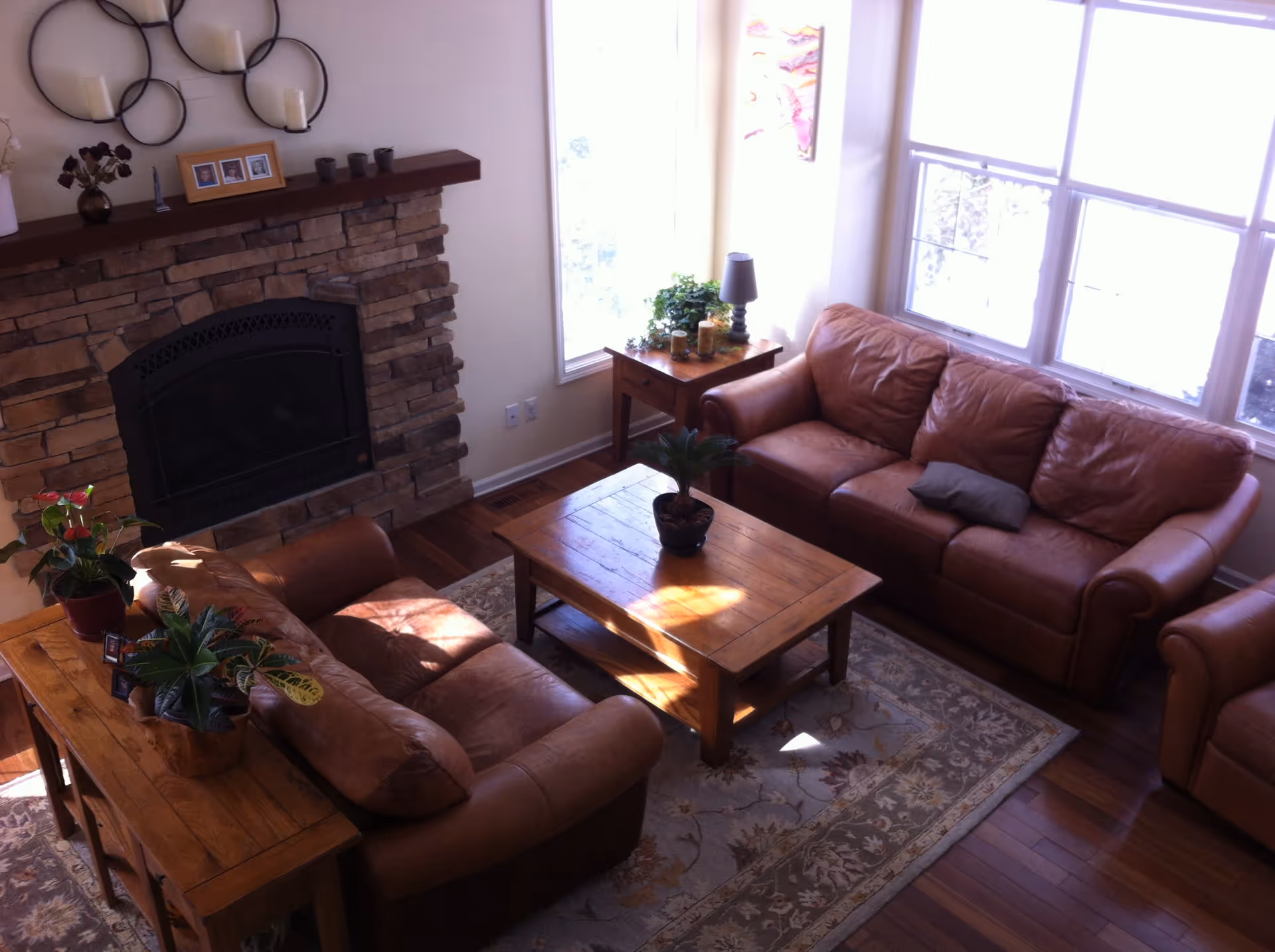 A cozy living room with two brown leather sofas, a wooden coffee table with a small plant on it, a side table with a lamp and decorative items, and a stone fireplace with candles and framed photos on the mantel. Large windows let in natural light, and a patterned rug covers the wooden floor.