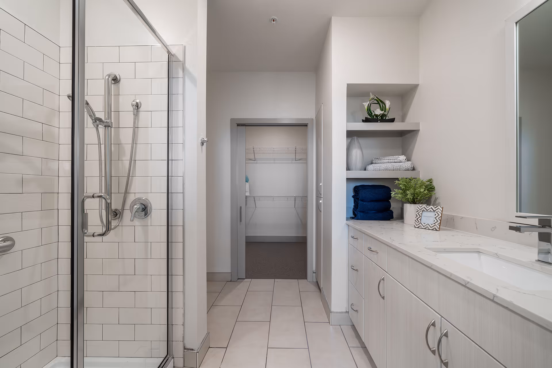 Bright modern bathroom featuring a glass-enclosed shower on the left, a long vanity with sink and open shelving on the right, and an open closet doorway straight ahead.