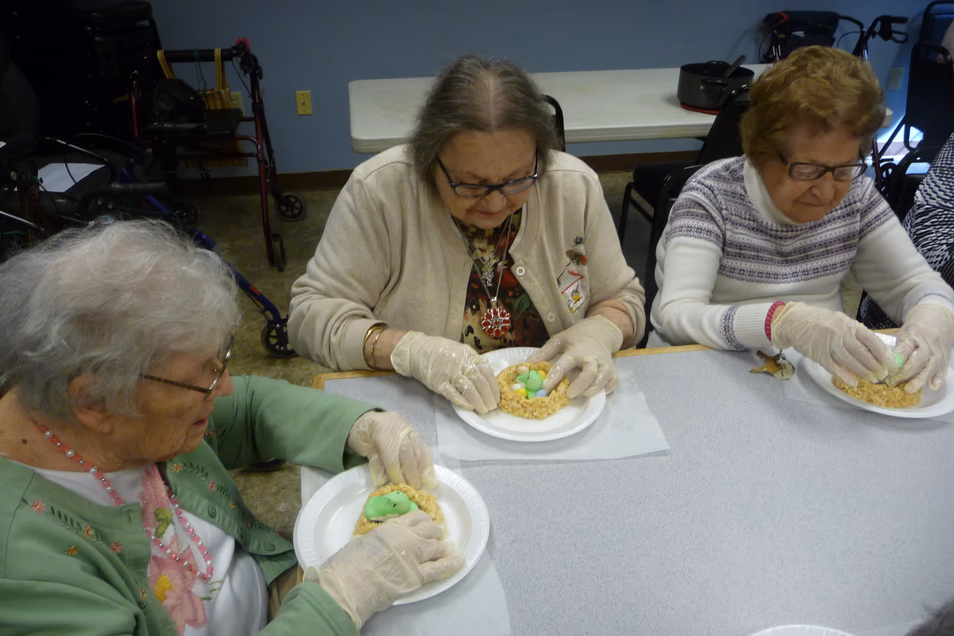 Three elderly women wearing gloves sit at a table in a room, each working on a craft or food project involving a circular base with colorful items on top, possibly candy or decorations. In the background, walkers and chairs are visible.