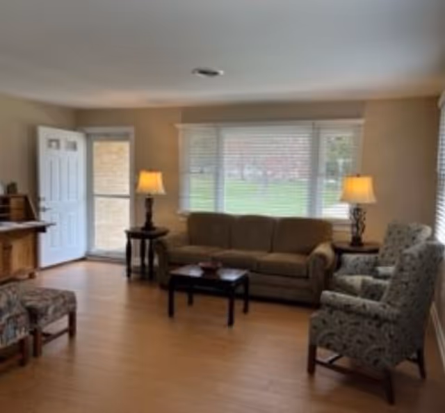 A cozy living room with a brown sofa, two patterned armchairs, a wooden coffee table, two side tables with lamps, a wooden desk, and a door open to the outside. Large windows with blinds allow natural light to fill the room.