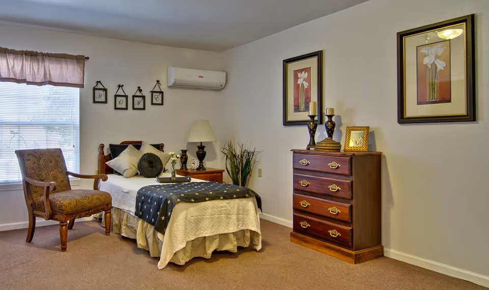 A cozy bedroom in a senior living facility featuring a single bed with white and dark blue bedding, decorative pillows, a wooden armchair with patterned upholstery, a wooden nightstand with a lamp, and a wooden dresser with decorative items and framed artwork on the walls.