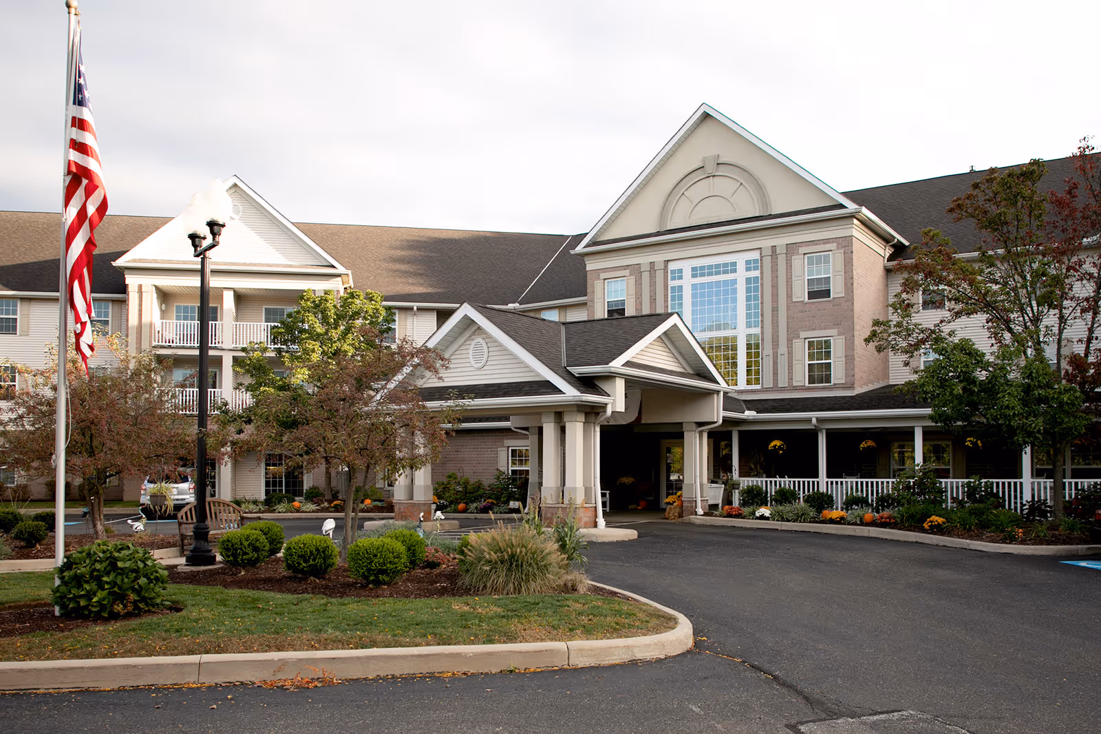 Exterior front view of a senior living facility building with a covered entrance, landscaped garden with bushes and trees, an American flag on a flagpole, and a paved driveway.