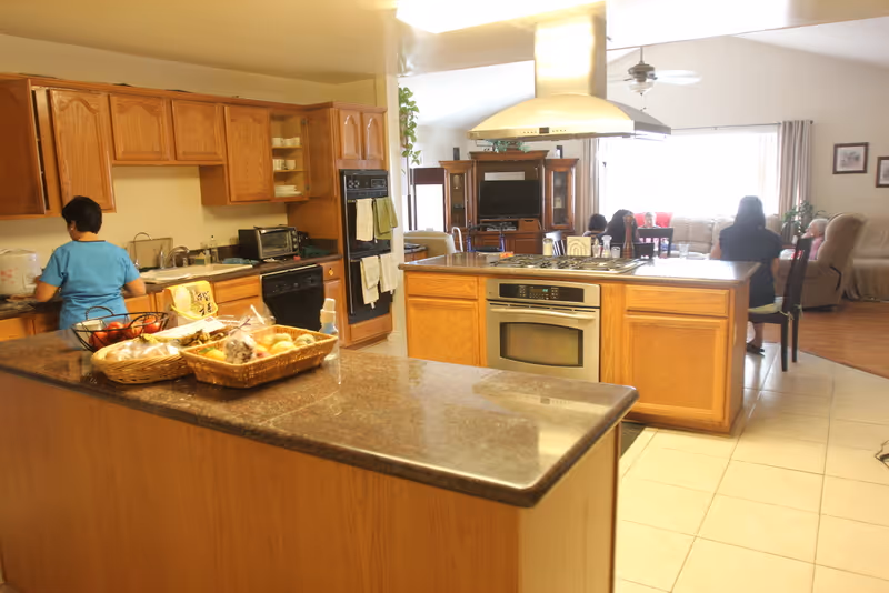 A spacious kitchen area with wooden cabinets and granite countertops. A person in a blue shirt is standing near the sink. The kitchen island has a built-in oven and stove with a range hood above it. In the background, there is a living room area with a television, chairs, and people sitting and conversing.