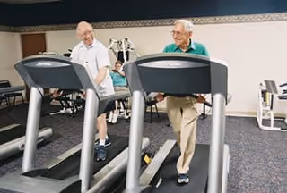 Two elderly men walking on treadmills in a fitness room, smiling and enjoying their exercise. In the background, a woman is seated on an exercise bike.
