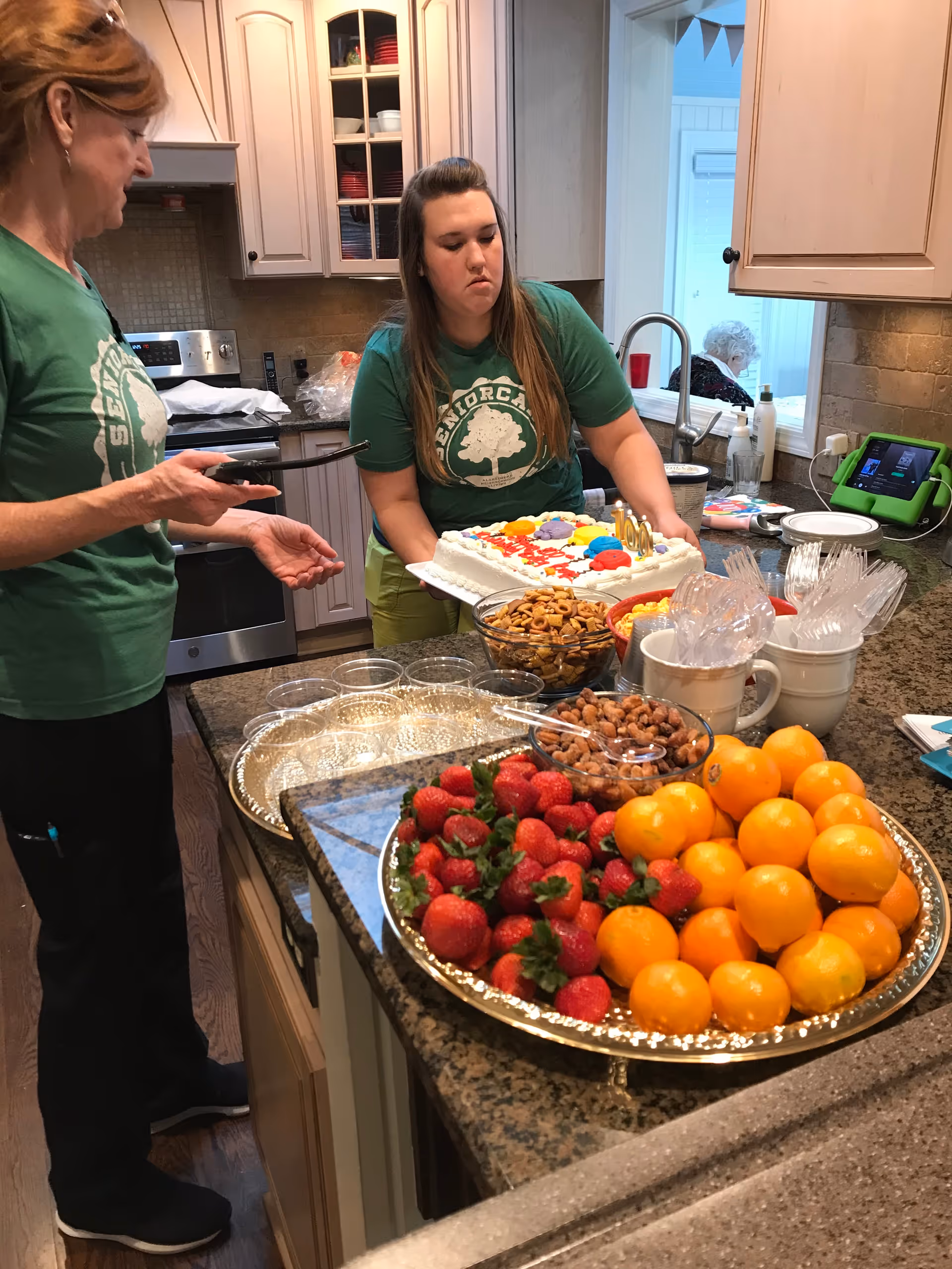 Two women in green SeniorCare Homes t-shirts are in a kitchen preparing for a celebration. One woman is holding a decorated cake with colorful icing, while the other is holding a serving utensil. On the kitchen counter, there are trays with strawberries, oranges, nuts, plastic cups, and utensils. In the background, an elderly person is seated near a window.