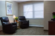 A cozy sitting area with two dark brown leather recliners positioned near a window with closed blinds. A small wooden side table with a lamp and a potted plant is placed between the chairs. The walls are light-colored with a framed picture hanging on one side. The floor is carpeted in a neutral tone.