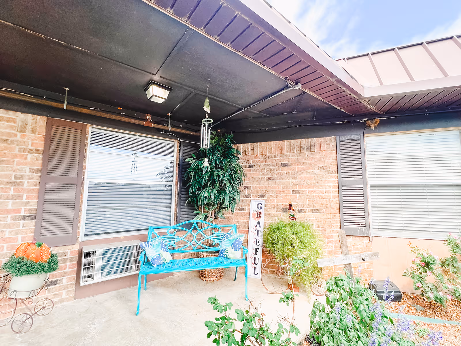 Covered outdoor patio area with a turquoise metal bench adorned with two decorative pillows, a large potted plant, a vertical sign that reads 'GRATEFUL', and various other plants and decorations including a small pumpkin in a planter and a wind chime hanging from the ceiling.