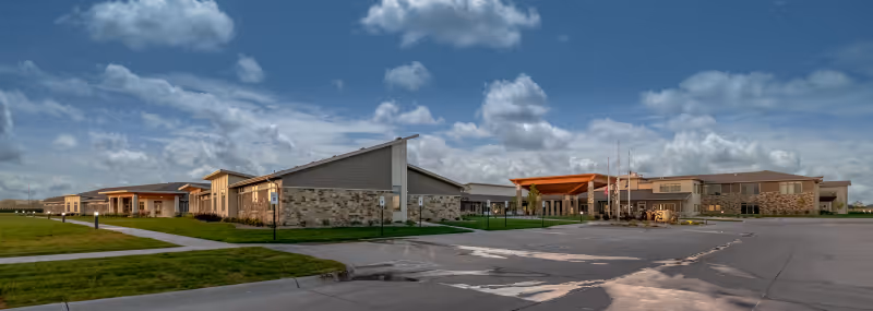 Panoramic exterior view of Brookestone Gardens senior living campus showing low-rise buildings, landscaped lawns, and a driveway under a cloudy sky.