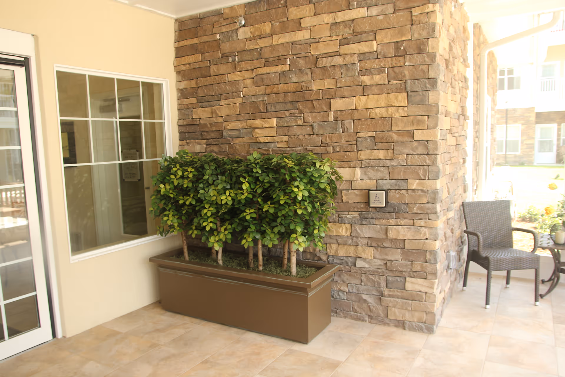 A covered outdoor patio area with a stone wall, a large rectangular planter with green leafy plants, a window, a glass door, and a wicker chair with a small round table holding a potted plant.
