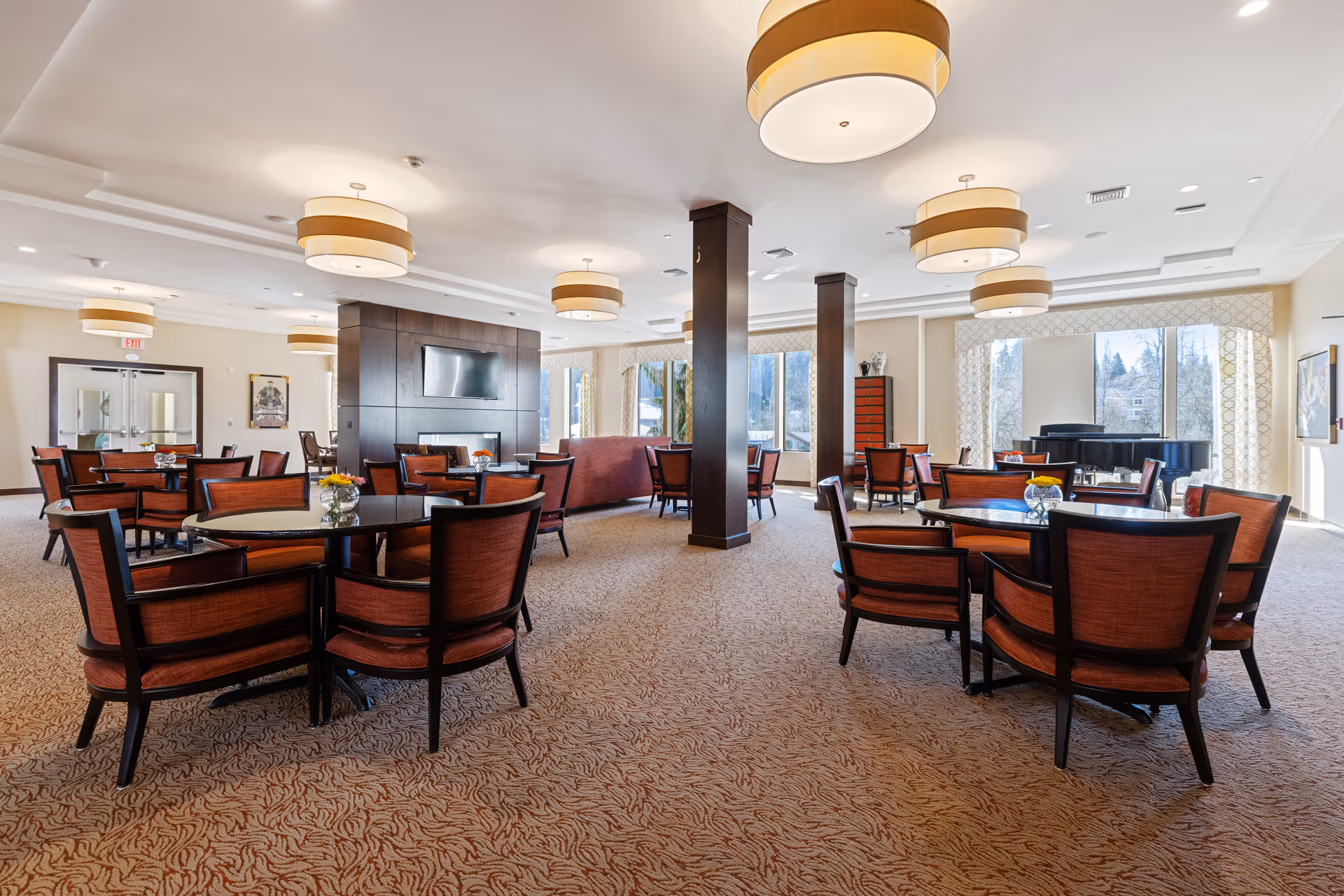 Bright communal dining and lounge area with round tables, upholstered chairs, a piano, and large windows.