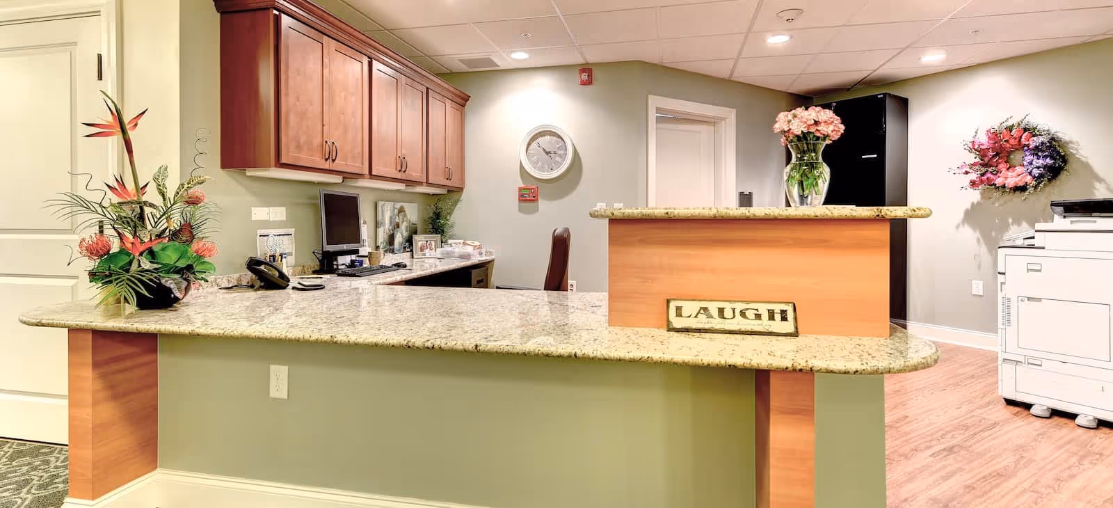 Reception desk area in a senior living facility with a granite countertop, wooden cabinets, a computer, a telephone, and decorative flower arrangements. A sign on the desk reads 'LAUGH'. The walls are light-colored, and there is a clock and a wreath on the walls.