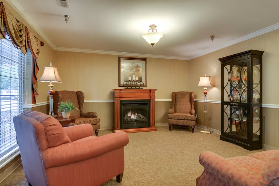 A cozy living room with beige walls and carpet, featuring a gas fireplace with a framed picture above it. There are two patterned armchairs and two pink upholstered chairs arranged around the fireplace. Two floor lamps with beige lampshades provide lighting. A black glass-front cabinet displaying decorative items is positioned against the right wall. A large window with blinds and draped curtains is on the left side of the room.