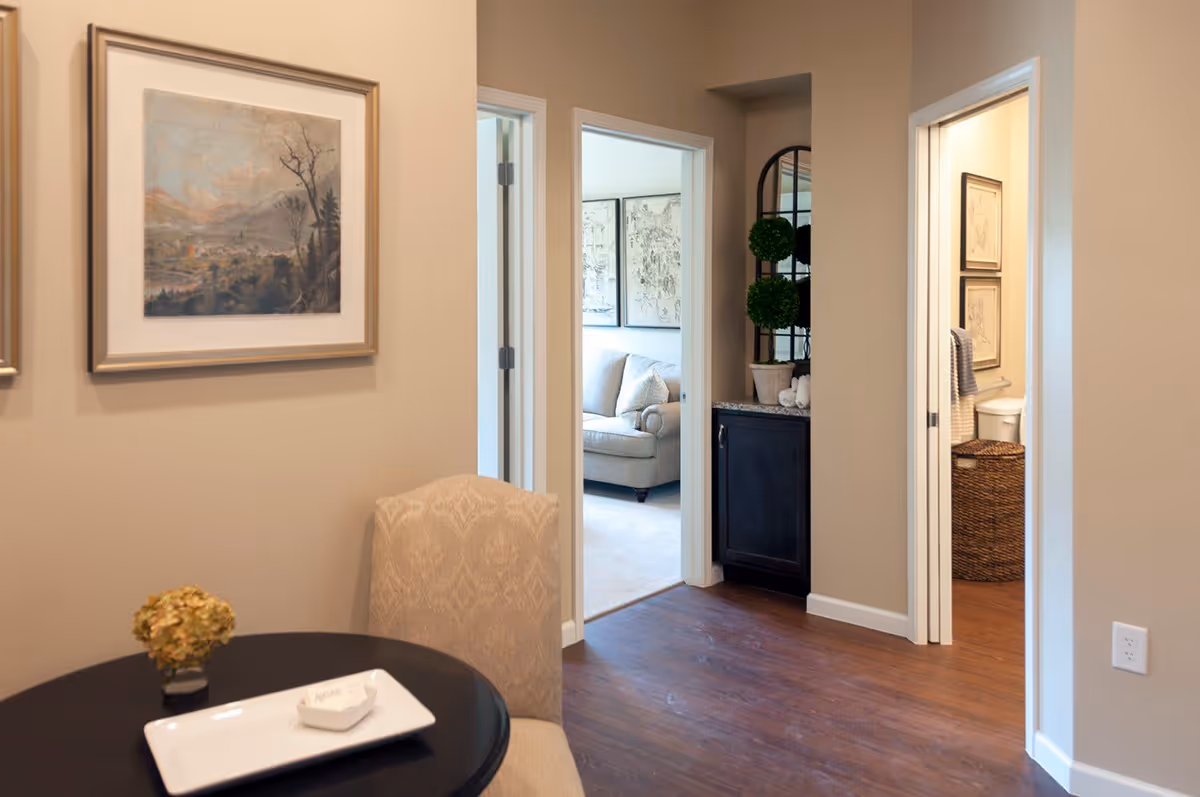 Interior view of a senior living facility showing a small round table with a chair in the foreground, two open doorways leading to a living room with a sofa and a bathroom with a toilet and framed pictures, beige walls, wooden floor, and decorative plants on a cabinet.