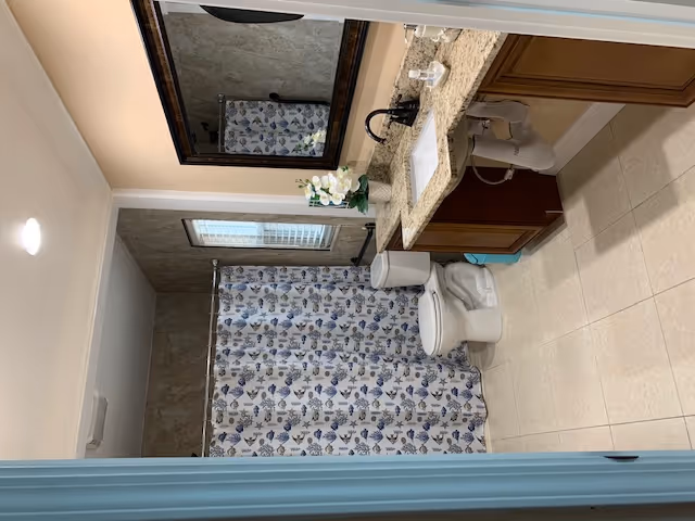 A clean bathroom with beige tiled floor and walls. There is a white toilet next to a bathtub with a shower curtain featuring a blue and beige seashell pattern. A granite countertop with a sink and dark faucet is on the right side, with a large framed mirror above it. A small potted plant with white flowers is placed on the countertop.