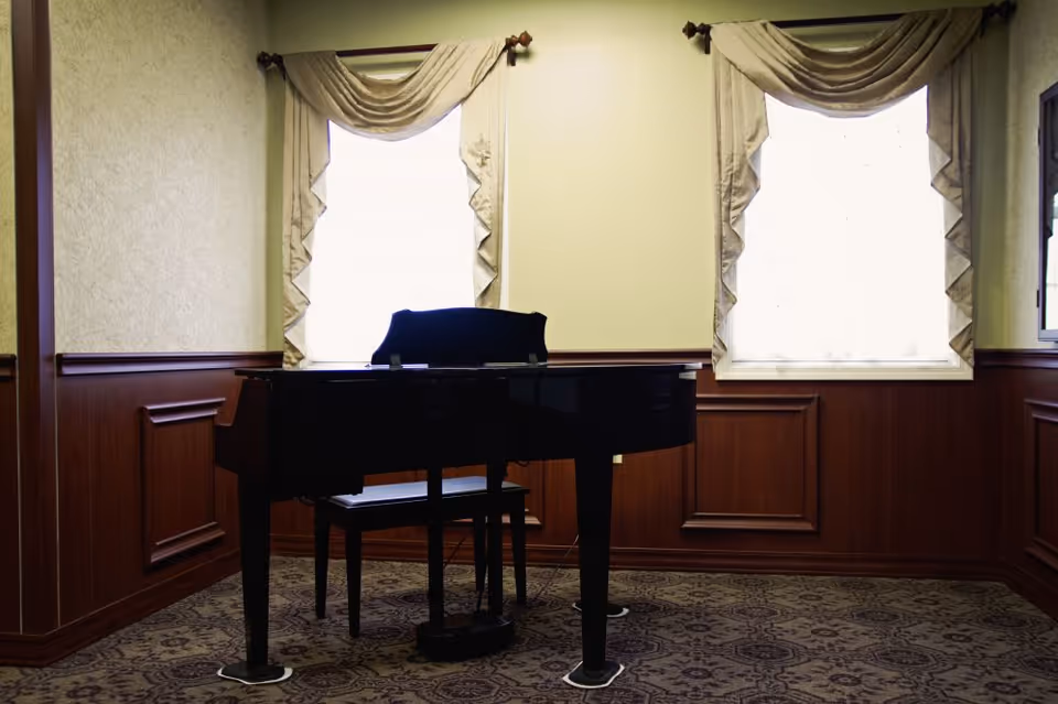 A black grand piano with a matching bench is placed in a room with two large windows covered by cream-colored drapes. The walls have wood paneling on the lower half and light-colored wallpaper on the upper half. The floor is carpeted with a patterned design.