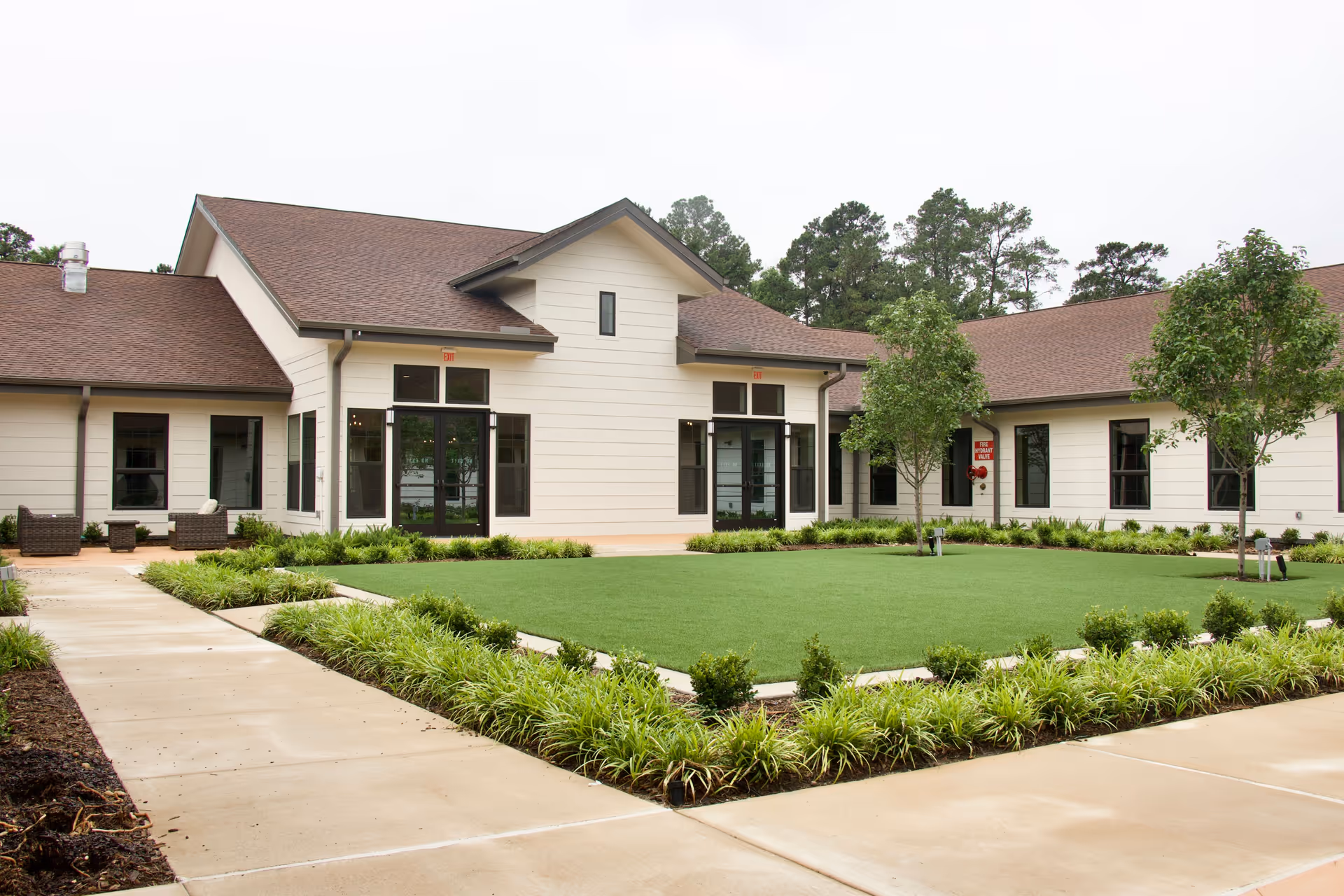 Exterior view of a single-story building with beige siding and brown roof shingles, surrounded by a well-maintained lawn and landscaped greenery. There are paved walkways leading to the building entrances, and a few small trees are planted around the lawn area.
