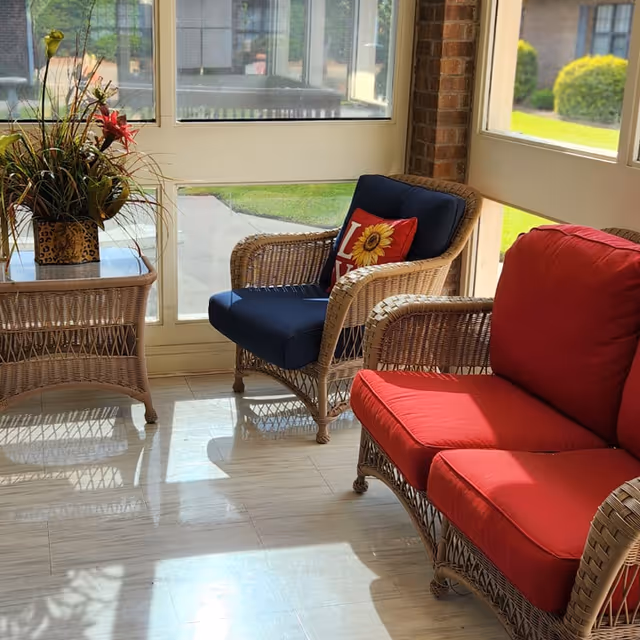 Sunlit seating area with wicker chairs featuring red and blue cushions, a wicker side table with a potted plant, and large windows overlooking a lawn.