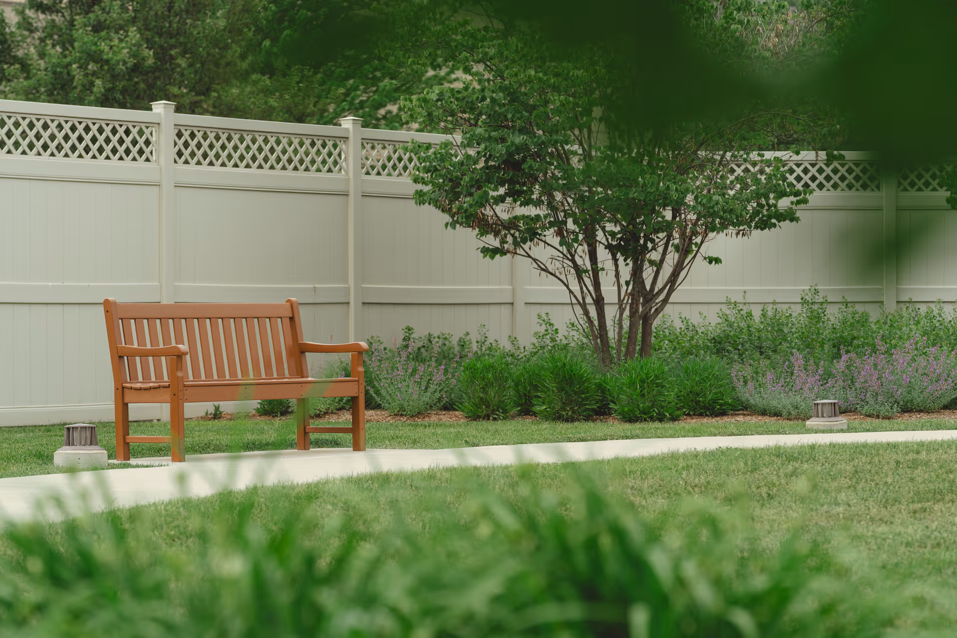 A wooden bench on a concrete path in a garden area with green grass, shrubs, a tree, and a white privacy fence in the background.