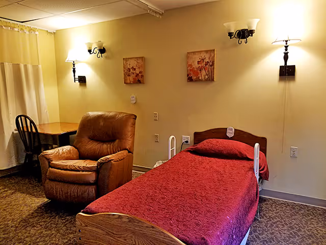 A cozy senior living bedroom featuring a single bed with a red bedspread, a brown cushioned armchair, a wooden desk with a black chair, two wall-mounted lamps, and two framed floral paintings on a beige wall.