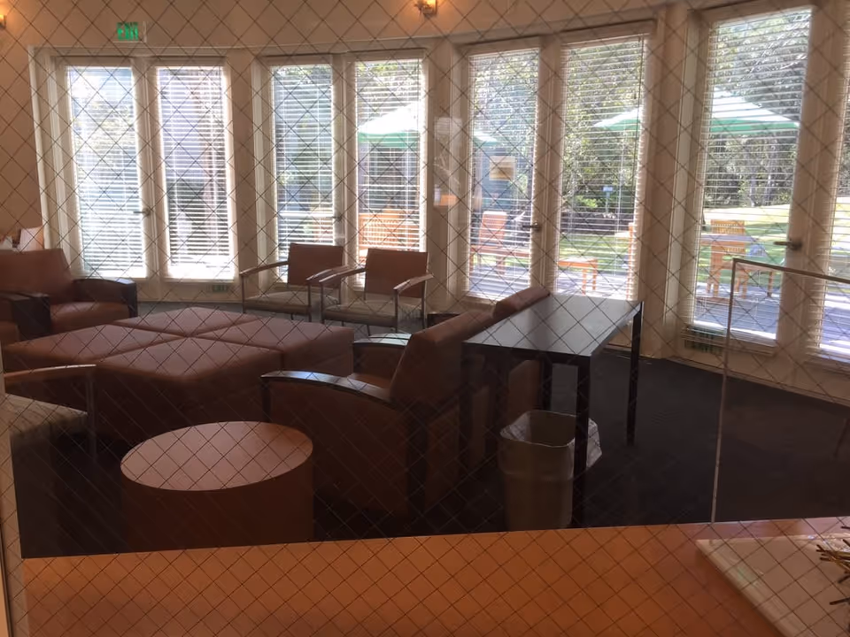 A lounge area with several armchairs, a round coffee table, and a rectangular table with a trash bin underneath. Large windows and glass doors with blinds reveal an outdoor patio with chairs and green umbrellas.