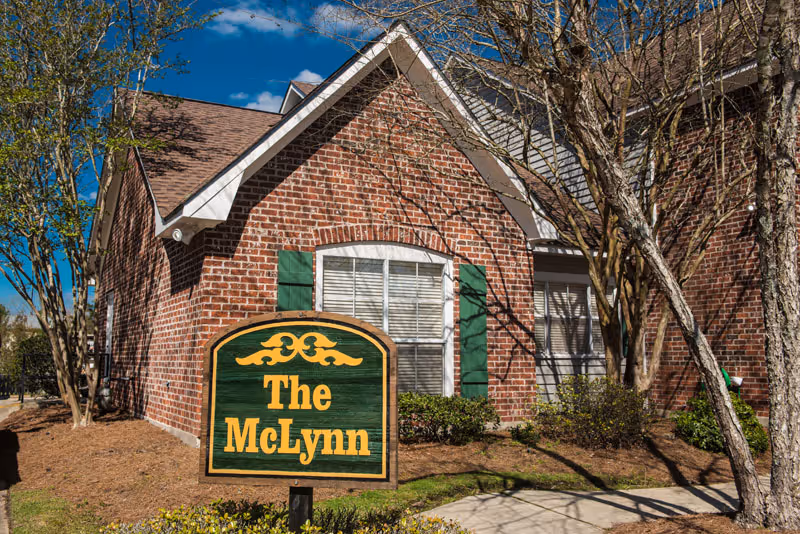 Exterior view of a brick building with green shutters and a sign in front that reads 'The McLynn'. There are trees and shrubs around the building under a clear blue sky.