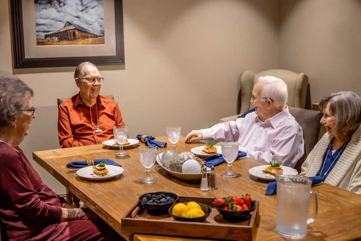 Four elderly people sitting around a wooden dining table, smiling and engaging in conversation. Each person has a plate with a small meal, blue napkins, and empty water glasses in front of them. On the table, there is a decorative bowl with spherical objects and a tray with bowls of blueberries, lemons, and strawberries. A pitcher of water is also visible. The room has beige walls and a framed picture of a barn hanging on the wall.