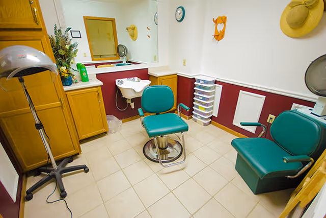 A small salon room with two green salon chairs, a hair dryer on a stand, a sink, wooden cabinets, a large mirror, and a stack of plastic drawers against a wall painted white and maroon. There are hats hanging on the wall and a clock above the drawers.