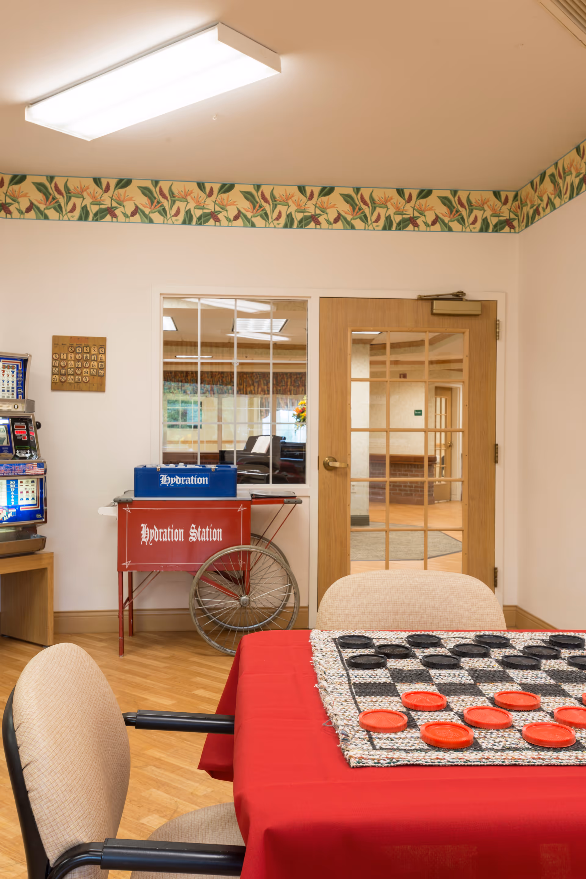 A cozy indoor common area with a table covered by a red tablecloth and a large checkers game set up with red and black pieces. Two beige chairs with black armrests are positioned around the table. In the background, there is a wooden door with glass panes and a window beside it. A red hydration station cart with a blue box labeled 'Hydration' sits against the wall. The room has a floral border near the ceiling and a wooden floor.