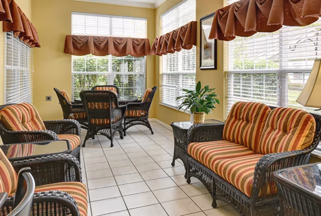 A sunlit sitting area with wicker furniture including a loveseat, chairs, and a table. The cushions have red and orange striped fabric. Large windows with white blinds and brown valances let in natural light. There is a small plant on a side table and a lamp in the corner.