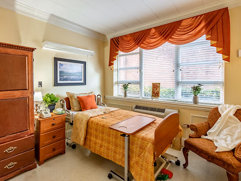 A cozy senior living bedroom with a single hospital-style bed covered in an orange and yellow patterned blanket. The bed has an adjustable overbed table and is positioned near a large window with white blinds and orange valance curtains. Next to the bed is a wooden nightstand with a telephone, lamp, and a small plant. There is a wooden armoire on the left side of the room and an upholstered armchair with a white blanket draped over it on the right side. A framed landscape painting hangs on the wall above the bed.
