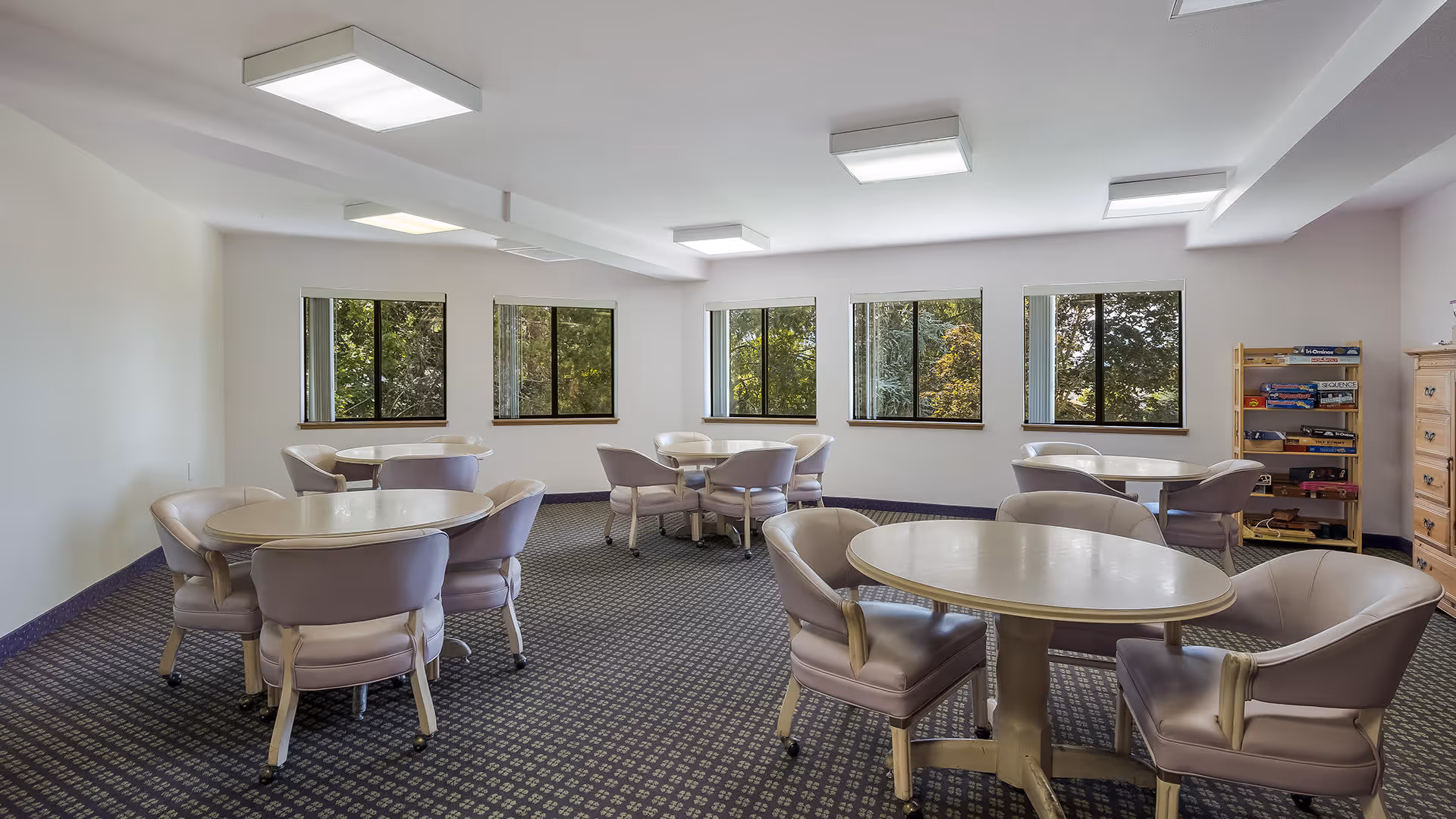 A bright room with multiple round tables, each surrounded by four cushioned chairs. The room has large windows showing green trees outside, a patterned carpet, and a wooden shelf with board games and puzzles in the corner.