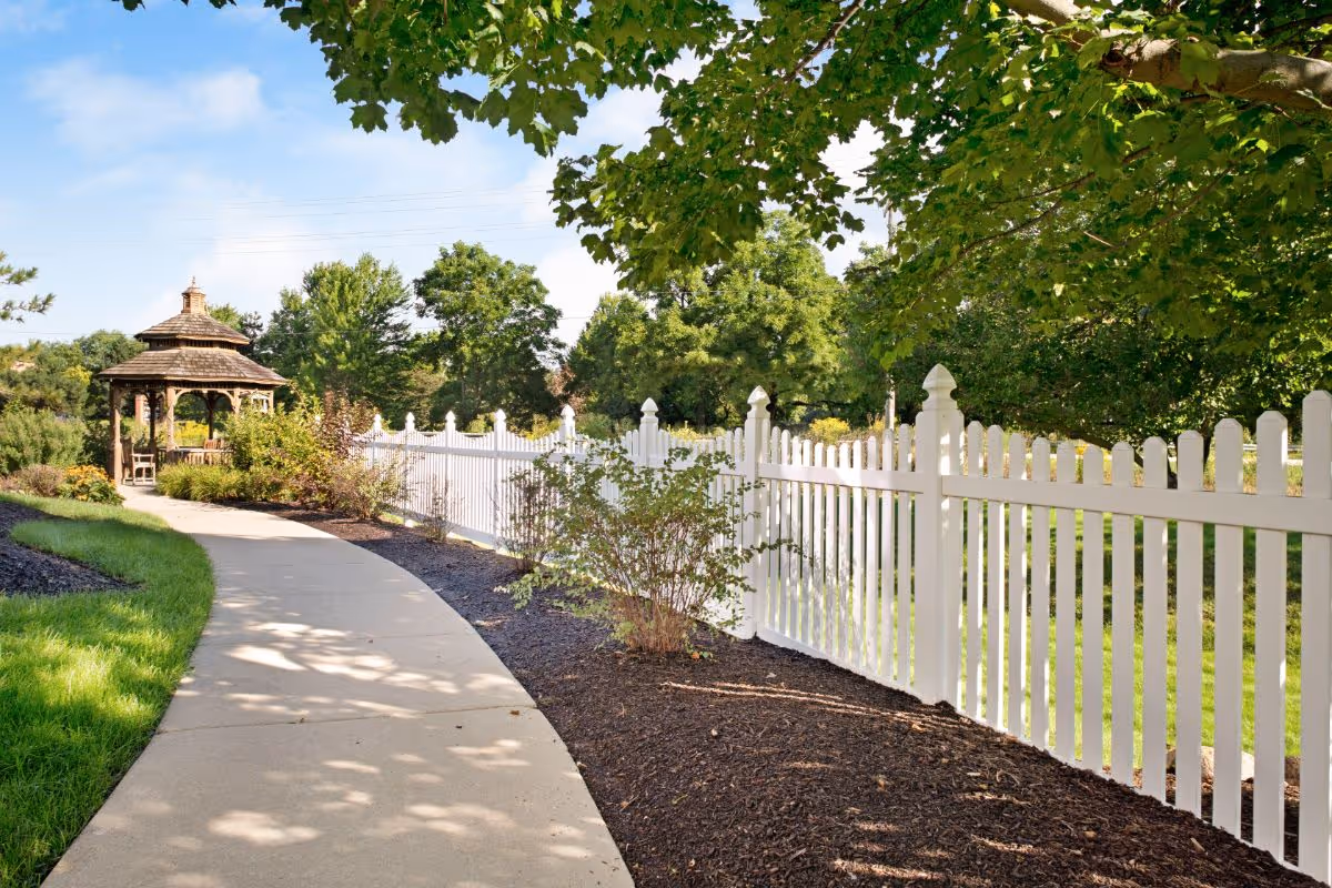 A curved concrete pathway bordered by green grass and mulch with small bushes, leading to a wooden gazebo surrounded by trees and a white picket fence under a blue sky with some clouds.