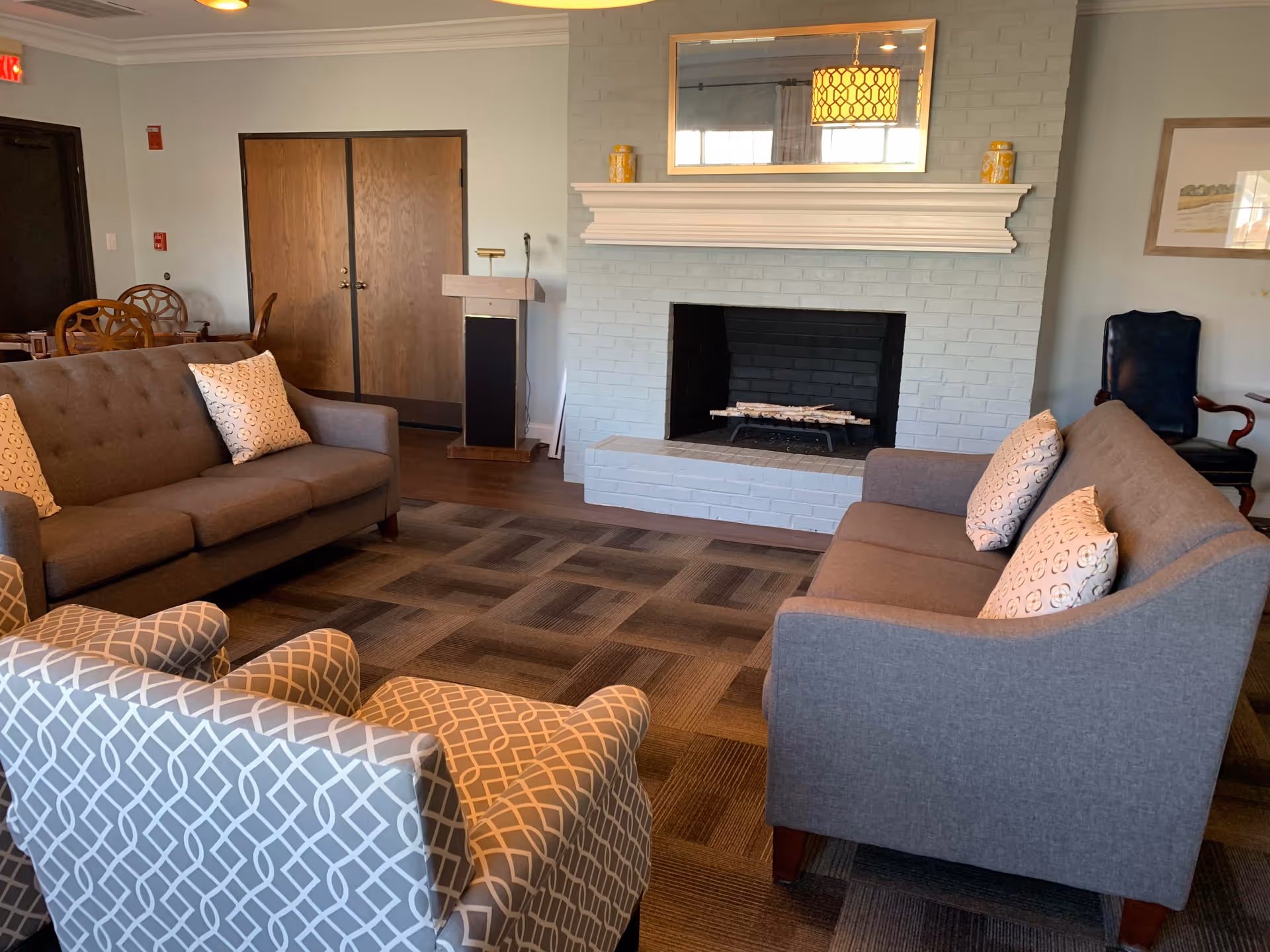 A cozy living room with two gray sofas and a patterned armchair arranged around a white brick fireplace. The fireplace has a white mantel with decorative items and a large mirror above it. There is a wooden podium and a set of double wooden doors in the background, along with a dining table and chairs partially visible.