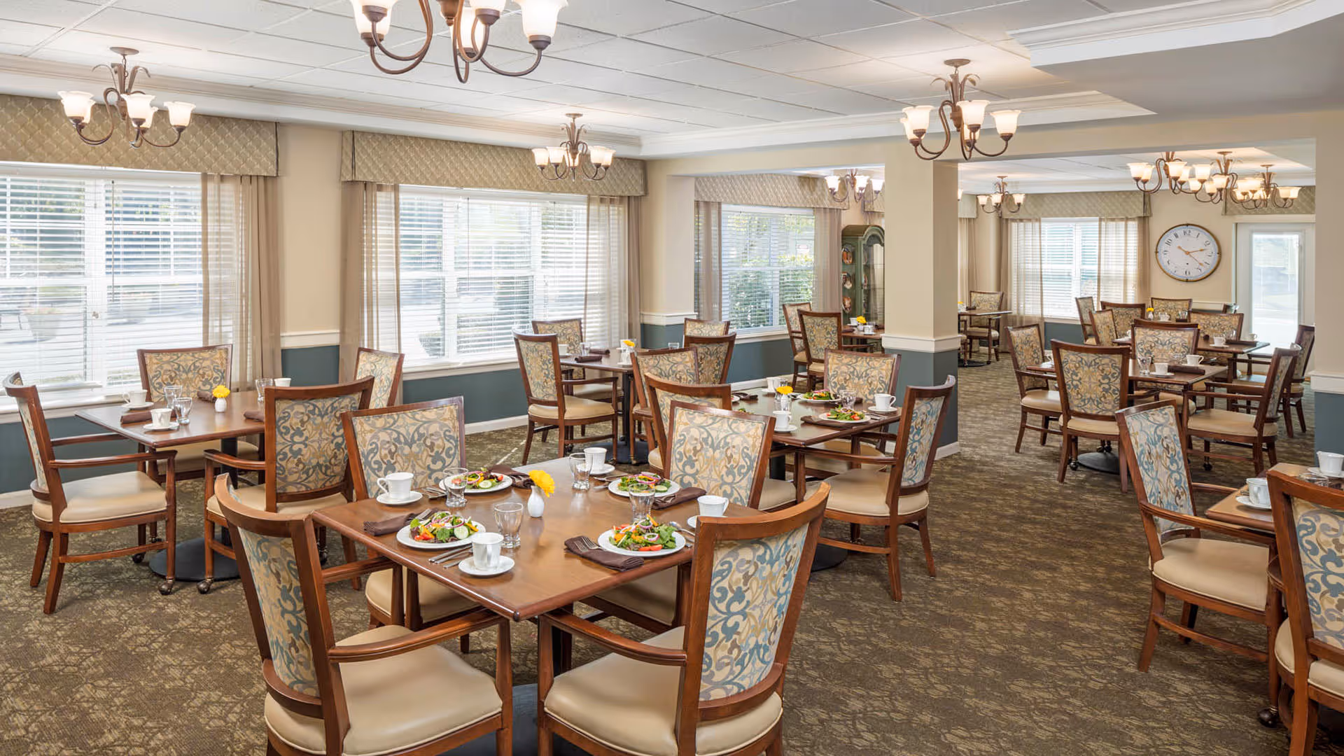 A bright and spacious dining room with multiple wooden tables and cushioned chairs arranged neatly. Each table is set with plates of salad, cups, glasses, and napkins. Large windows with blinds and valances allow natural light to fill the room. Elegant chandeliers hang from the ceiling, and a large clock is visible on the far wall.