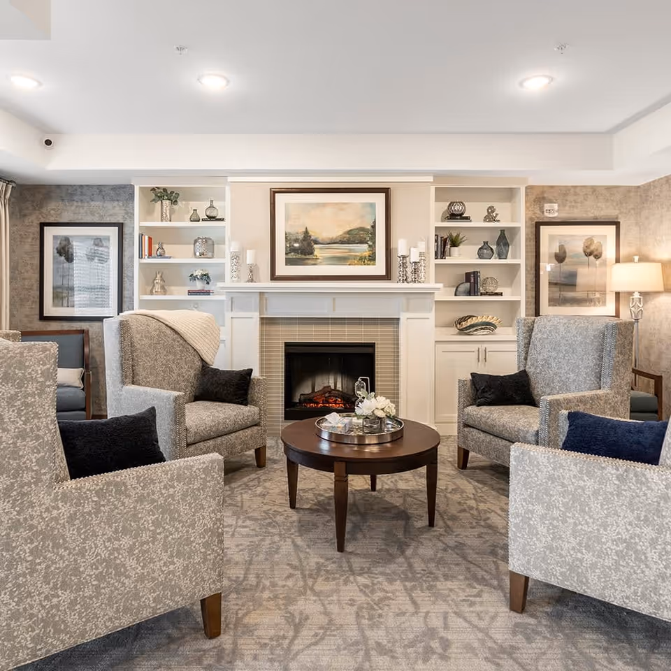 A cozy senior living room with four patterned armchairs arranged around a round wooden coffee table. The room features a fireplace with a landscape painting above it, built-in white shelves with decorative items, and framed artwork on the walls. Soft lighting and a carpeted floor add to the warm ambiance.