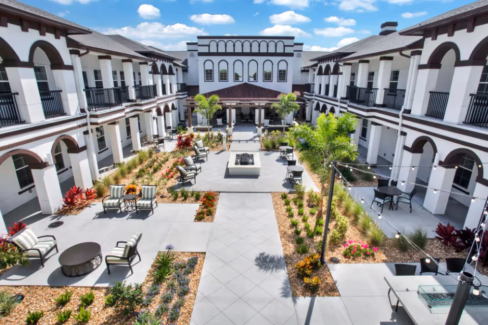 Sunny central courtyard of a two-story senior living facility with patio seating, landscaping, string lights, and a central fire pit.