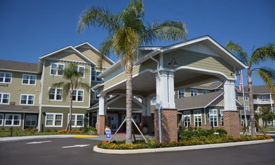 Exterior view of Orchard Heights Gracious Retirement Living facility showing a covered entrance with brick columns, palm trees, and a multi-story building with many windows under a clear blue sky.