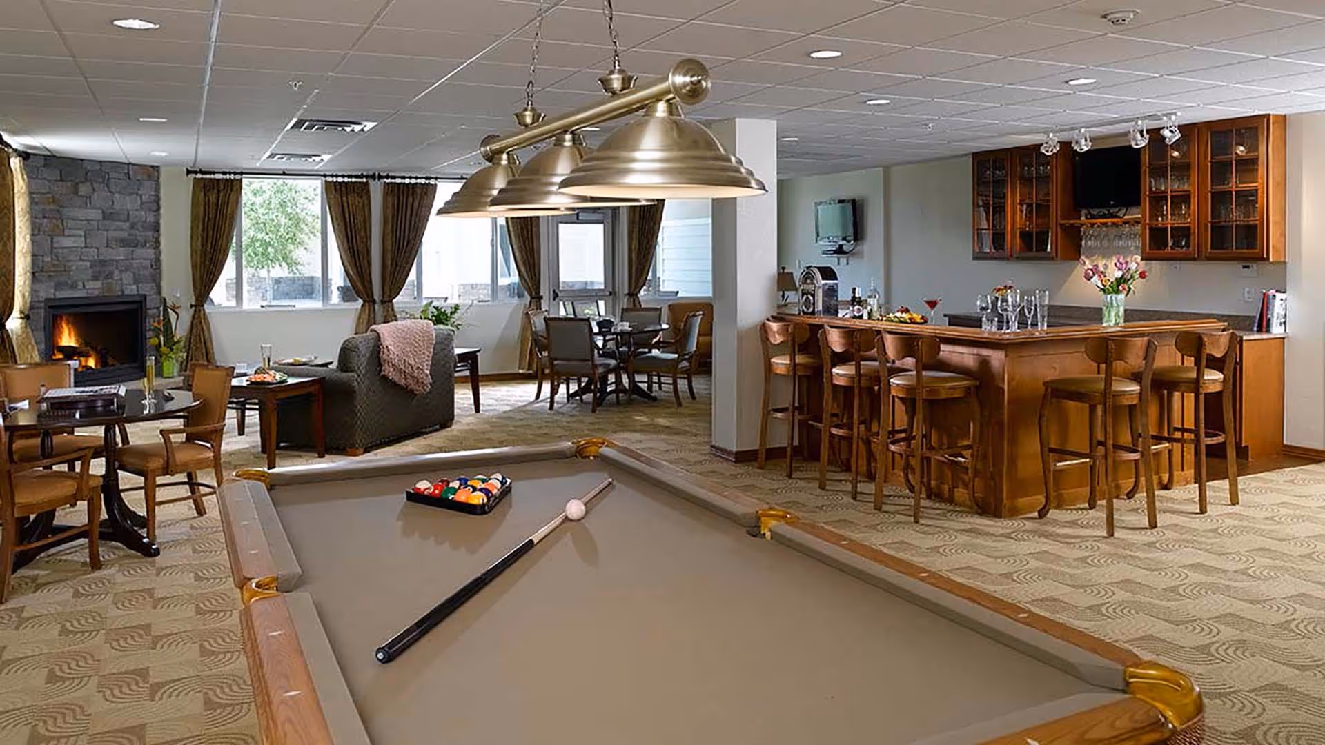 A spacious common area featuring a pool table with balls and cue stick, a wooden bar with stools, a cozy seating area with chairs and a sofa near a stone fireplace, and large windows with curtains letting in natural light.