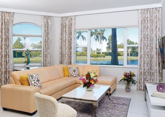 A bright living room with a large beige sectional sofa adorned with colorful throw pillows, a white coffee table with a floral arrangement, a patterned area rug, and large windows showing a view of palm trees and a lake outside. There are patterned curtains on the windows and a TV mounted on the wall to the right.
